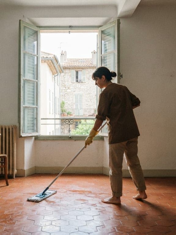 Femme en tunique brune passant une serpillière sur des tomettes devant une haute fenêtre à persiennes ouvertes